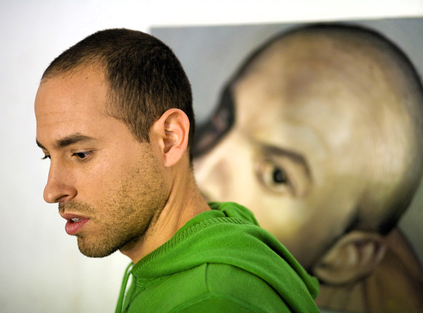 Artist Roi Pajursky seen from behind, working in his bright art studio. He is focused on a detailed portrait painting on an easel, surrounded by art supplies, brushes, and several framed fragments on the walls, capturing an authentic moment of the creative process.