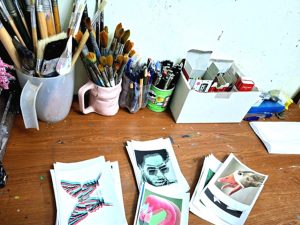 Artist Roi Pajursky workspace featuring art supplies and sketches on a wooden table. Hand-drawn illustrations and colorful drafts are displayed next to jars of paintbrushes, capturing the alchemy of materials and the creative process behind his kinetic artwork.