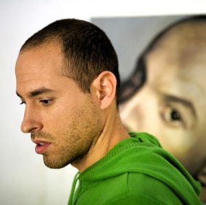Roi Pajursky - Artist portrait in his creative studio, sitting at his work desk surrounded by oil paints, brushes, and colorful surreal canvases, showing the authentic artistic process.
